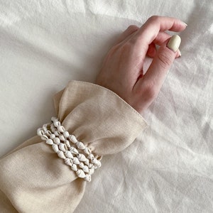 Può includere: Un braccialetto di conchiglie bianche con più file di piccole conchiglie bianche. Il braccialetto è indossato su una manica di maglione beige.