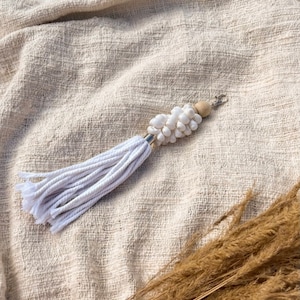 Op de afbeelding: Sleutelhanger van witte schelpen en houten kralen met een witte kwast op een beige stoffen achtergrond.