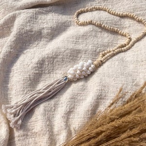 Può includere: Una collana di conchiglie bianche e perline di legno con un fiocco bianco. La collana è appoggiata su uno sfondo di tessuto bianco con erba secca beige.