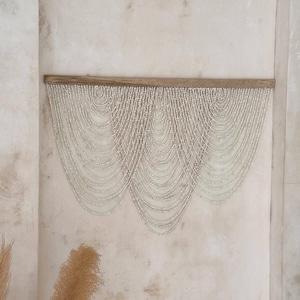 Op de afbeelding: Een wandhanger met een horizontale houten balk waaraan meerdere strengen kleine, gebroken witte kralen hangen. De kralen zijn gerangschikt in een reeks overlappende, trapsgewijze U-vormige slingers die een gelaagd, gedrapeerd effect creëren.