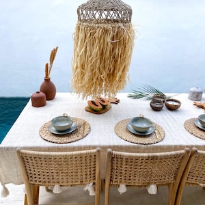 May include: A dining table setting with a woven rattan lampshade hanging above, two woven rattan chairs, and two place settings with woven placemats and bowls. The table is covered with a white tablecloth with tassels.