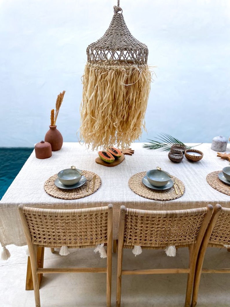 May include: A dining table setting with a woven rattan lampshade hanging above, two woven rattan chairs, and two place settings with woven placemats and bowls. The table is covered with a white tablecloth with tassels.