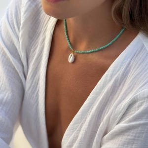 May include: A delicate necklace featuring small turquoise beads and a white seashell pendant. The necklace is worn against a white top, creating a contrast that highlights the jewelry's natural colors and textures.