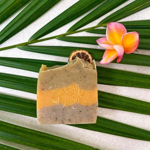 Pode incluir: Uma barra de sabão verde, laranja e marrom com o texto "bali bali" nela. O sabão está em cima de uma folha de palmeira verde.