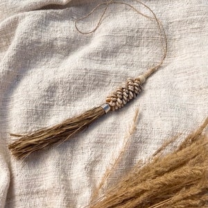 May include: A white and brown tassel with a shell and bead design hanging on a white fabric background.