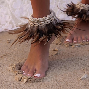 May include: A pair of ankle bracelets featuring brown feathers and white seashells. The bracelets are worn on a person's ankles, and the person is standing on a sandy beach.