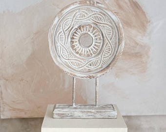 Puede incluir: Una escultura decorativa de madera blanca con un diseño circular, montada sobre un soporte metálico y una base cuadrada blanca. El diseño circular presenta intrincadas tallas. La escultura se exhibe sobre un fondo neutro.