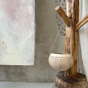 Pode incluir: Um vaso pendurado feito de materiais naturais com um design delicado e intrincado. O vaso est&aacute; pendurado em um suporte de madeira r&uacute;stico com uma pe&ccedil;a de rede.