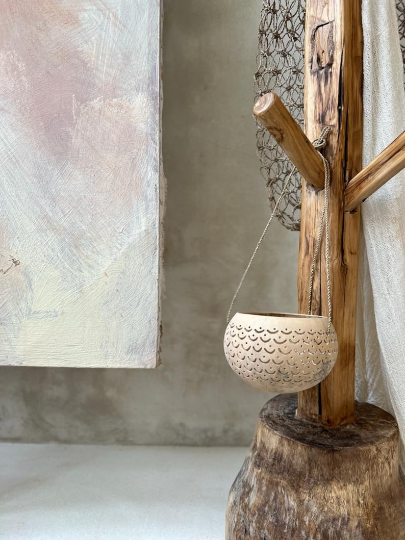 Pode incluir: Um vaso pendurado feito de materiais naturais com um design delicado e intrincado. O vaso est&aacute; pendurado em um suporte de madeira r&uacute;stico com uma pe&ccedil;a de rede.