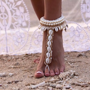 Peut inclure: Un bracelet de cheville en perles blanches et dorées avec des coquillages. Le bracelet est porté sur le pied d'une femme sur une plage de sable.
