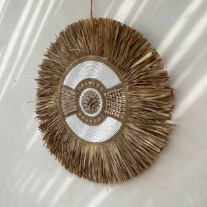 Könnte beinhalten: Ein runder Wandbehang aus natürlichen Materialien mit einem gewebten Design mit weißen Perlen und einem Fransen aus braunen Fasern.
