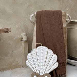 May include: A white seashell-shaped decorative item with a textured surface and a brown rope base. A brown textile is draped over a large, light-colored vase. The background features a neutral-toned wall and floor.