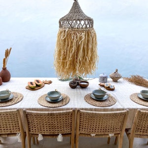May include: A table setting with woven placemats, bowls, and silverware. The table is covered in a white linen tablecloth. A woven pendant light hangs above the table.