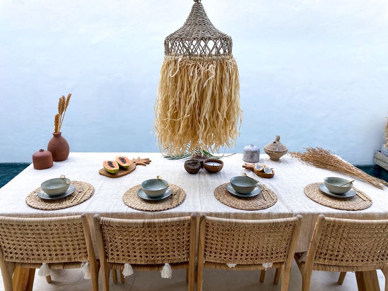 May include: A table setting with woven placemats, bowls, and silverware. The table is covered in a white linen tablecloth. A woven pendant light hangs above the table.