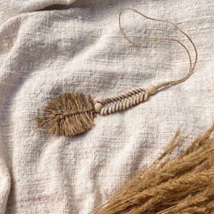Könnte beinhalten: Eine handgefertigte Halskette mit einem natürlichen, böhmischen Design. Die Halskette hat einen gewebten Raffia-Anhänger, eine Kette aus kleinen, weißen Muscheln und eine Holzperle. Die Halskette wird auf einem strukturierten, cremefarbenen Stoff präsentiert.