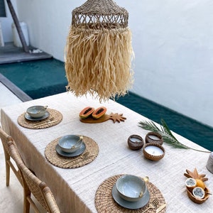 May include: A table setting with woven placemats, blue bowls, and gold silverware. The table is set for four people and is decorated with a woven lampshade, a pineapple, and a papaya. The table is set on a patio with a pool in the background.