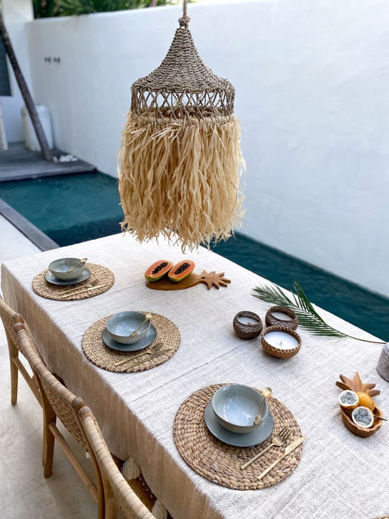 May include: A table setting with woven placemats, blue bowls, and gold silverware. The table is set for four people and is decorated with a woven lampshade, a pineapple, and a papaya. The table is set on a patio with a pool in the background.