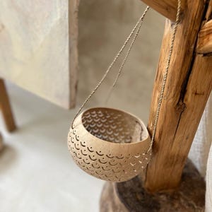 Pode incluir: Um vaso pendurado feito de uma casca de coco natural com um padr&atilde;o de corte decorativo. O vaso est&aacute; suspenso de uma viga de madeira com uma corda fina.