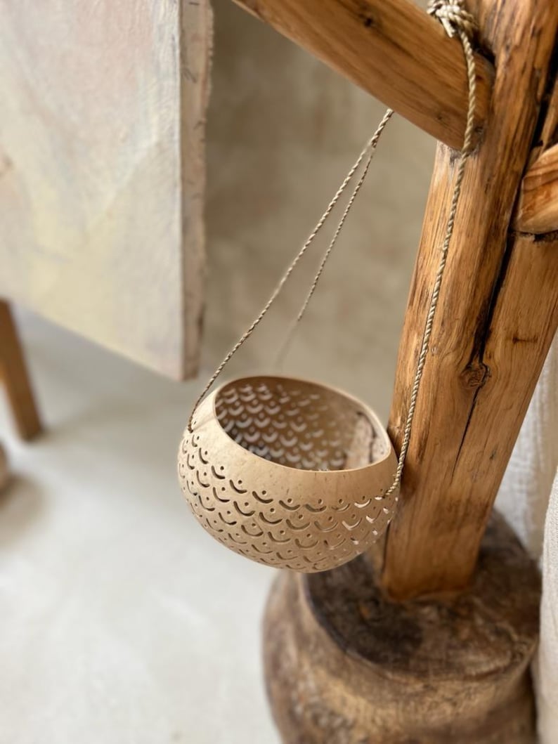 Pode incluir: Um vaso pendurado feito de uma casca de coco natural com um padr&atilde;o de corte decorativo. O vaso est&aacute; suspenso de uma viga de madeira com uma corda fina.