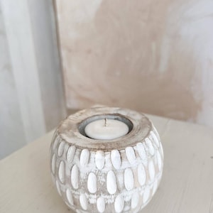May include: A round, white-washed wooden candle holder with a carved oval pattern. The candle holder has a small metal insert to hold a tea light candle. The candle is white and unlit.