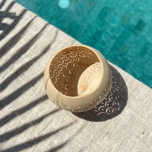 Pode incluir: Uma tigela de casca de coco branca com intrincados desenhos recortados, apresentando um padr&atilde;o de lua crescente. A tigela est&aacute; sentada sobre uma superf&iacute;cie texturizada branca com sombras de uma palmeira.
