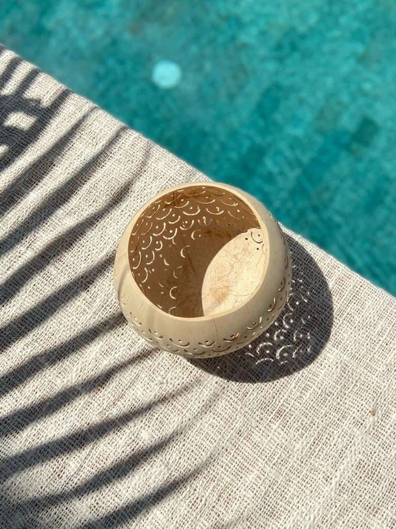 Pode incluir: Uma tigela de casca de coco branca com intrincados desenhos recortados, apresentando um padr&atilde;o de lua crescente. A tigela est&aacute; sentada sobre uma superf&iacute;cie texturizada branca com sombras de uma palmeira.