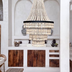 Könnte beinhalten: Ein mehrstufiger Kronleuchter mit einem schwarzen Metallrahmen und Strängen aus hellfarbigen Holzperlen. Der Kronleuchter hängt in einem Raum mit weißen Wänden und Holzschränken und schafft eine warme Atmosphäre.