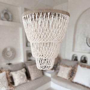 May include: A white shell chandelier with a natural jute rope trim. The chandelier has three tiers of shells and is hanging from the ceiling in a room with a white sofa and cushions.