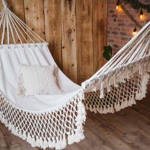 May include: A white cotton hammock with fringe detailing hangs in a rustic setting. The hammock is made of a soft, woven fabric and has a pillow for added comfort.