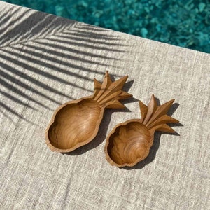 May include: Two wooden pineapple-shaped bowls. The bowls are made of light brown wood and have a carved design. The bowls are on a beige linen surface. The background includes a pool of water.