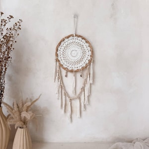 May include: A handmade dreamcatcher with a woven brown ring and a white, crocheted centre. It features hanging white tassels, wooden beads, and small shell accents. The dreamcatcher is suspended against a neutral-toned wall, showcasing a bohemian aesthetic.