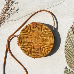 May include: A round, woven straw bag with a brown leather strap. The bag has a natural, light brown color with a slightly darker brown woven pattern.