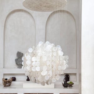 May include: A white, spherical chandelier composed of numerous circular, shell-like elements hangs below a large, circular, textured ceiling fixture. The light fixtures are suspended in a room with arched wall features and wooden cabinetry.