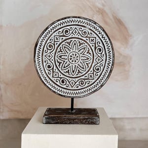 May include: A circular wooden ornament featuring a white, carved floral pattern. It is mounted on a black metal stand and a rectangular wooden base. The colour scheme is brown and white, set against a neutral background.