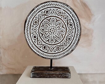 Puede incluir: Una pieza decorativa redonda de madera con un diseño floral blanco tallado. Está montada sobre un soporte de metal negro y una base rectangular de madera. La combinación de colores es marrón y blanco, con un fondo neutro.