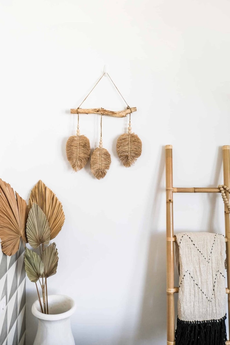 K&ouml;nnte beinhalten: Ein Wandbehang aus nat&uuml;rlichen Materialien mit drei gewebten Blattformen, die an einem Ast h&auml;ngen. Der Ast ist mit einem wei&szlig;en Haken an der Wand befestigt. Der Wandbehang ist hellbraun und hat einen rustikalen, bohemian Stil.