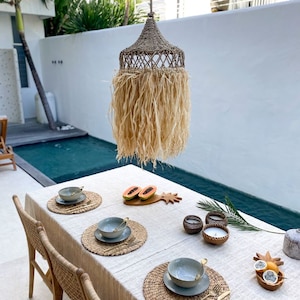 May include: A tropical-themed dining table setting with woven placemats, gray bowls, and gold cutlery. The table is set with papaya slices on a wooden cutting board, candles in wooden holders, and a pineapple-shaped serving dish. The table is set against a backdrop of a swimming pool and a woven pendant light.
