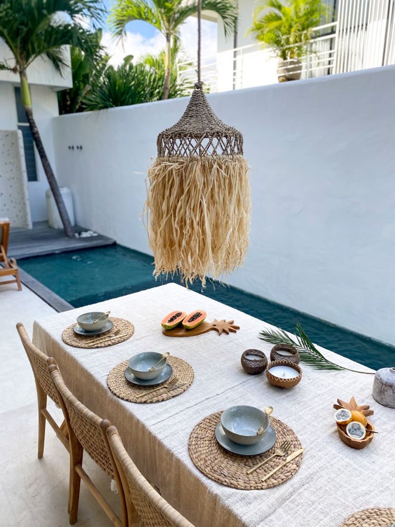May include: A tropical-themed dining table setting with woven placemats, gray bowls, and gold cutlery. The table is set with papaya slices on a wooden cutting board, candles in wooden holders, and a pineapple-shaped serving dish. The table is set against a backdrop of a swimming pool and a woven pendant light.