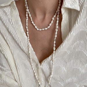 Puede incluir: Un collar largo de conchas blancas con un diseño delicado y en capas. El collar está hecho de pequeñas conchas blancas que están enhebradas en un cordón delgado. El collar lo lleva una persona con una camisa blanca.