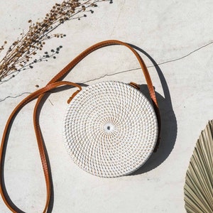 Könnte beinhalten: Eine weiße runde geflochtene Tasche mit einem braunen Lederriemen. Die Tasche liegt auf einer hellgrauen Oberfläche mit getrockneten Blumen und Palmblättern.