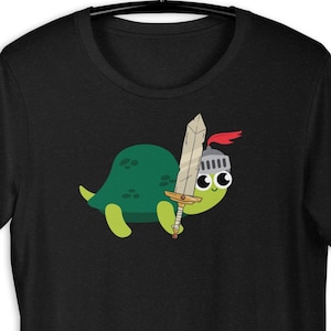 Op de afbeelding: Zwart t-shirt met een cartoon schildpad ridder ontwerp. De groene schildpad draagt een zilveren helm met een rode pluim en houdt een zwaard vast met een gouden handvat en zilveren lemmet. Het ontwerp staat op de voorkant.
