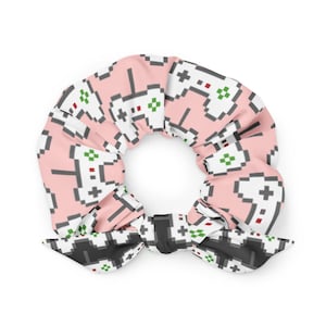 May include: Pink scrunchie with a pixelated video game controller pattern in white, gray, red, and green. The scrunchie has a bow detail with a black and white controller pattern. A fun accessory for gamers.