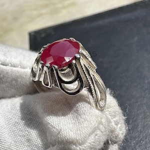 May include: A silver ring featuring an oval, deep red gemstone. The ring has an ornate, detailed design with curved and textured elements around the stone and on the band. The ring is displayed on a white surface.