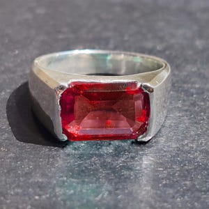 May include: A silver ring with a rectangular, deep red gemstone. The gemstone is set in a simple, classic band. The ring is displayed on a dark, textured surface, highlighting the vibrant color of the stone.