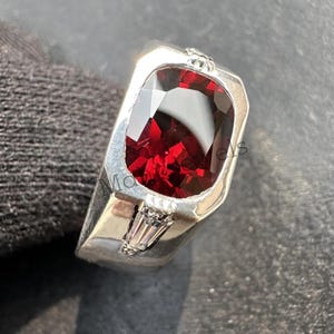 May include: A silver ring featuring a large, rectangular, deep red gemstone. The ring has a modern design with a rectangular setting and small, clear accent stones. The band is wide and polished, creating a striking contrast with the dark gemstone.