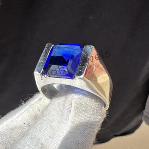 May include: A silver ring featuring a large, rectangular, deep blue gemstone. The ring has a modern design with a thick band and a setting that highlights the vibrant color of the stone. The ring is held by a gloved hand.