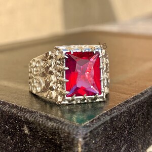 May include: Silver-toned ring featuring a large, rectangular, vibrant red gemstone. The ring has a detailed, floral-like pattern on the band and around the gemstone setting. The gemstone is the focal point, catching the light and providing a striking contrast to the silver metal.