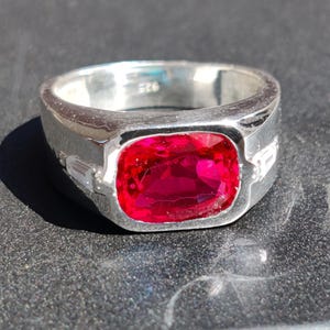 May include: A silver ring with a large, rectangular, deep red gemstone. The ring has a wide band and small, rectangular accent stones on each side of the main stone. The metal is stamped with "925".