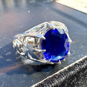 May include: A silver ring with a large, oval-cut, deep blue gemstone. The ring has an ornate, openwork design on the band, featuring stylized leaf or floral patterns. The gemstone is the focal point, catching the light and displaying a brilliant shine.