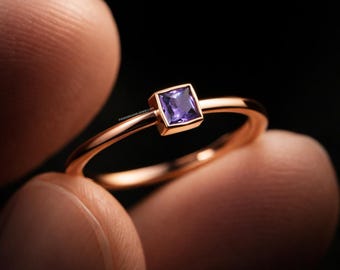 May include: A rose gold ring with a square, purple gemstone. The ring has a thin band and a bezel setting for the gemstone. The ring is held up against a dark background.